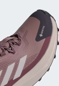 Mauve spalvos GORE-TEX bėgimo bateliai su tekstūruota viršutine dalimi, sustiprinta nosimi ir trimis baltomis juostelėmis. Pasižymi tvirtu padu su sukibimu.