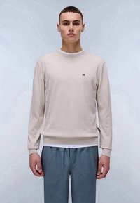 Sweatshirt de malha bege claro com mangas longas, punhos e bainha canelados. Pequeno logotipo vermelho no peito. Combinado com calças cinzas.