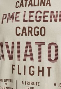 T-shirt en coton crème avec un large texte bordeaux foncé et des accents rayés. Le texte comprend "PME Legend", "Cargo" et "Aviation Flight."
