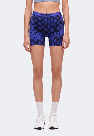 Femme portant un short de sport Jordan bleu et noir à motifs, des chaussettes blanches montantes et des baskets Nike blanches, debout devant un fond uni.