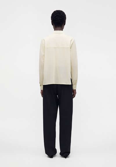 Personne debout de dos, portant une chemise crème à manches longues et un pantalon noir ample sur un fond blanc uni.