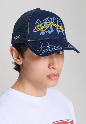Joven con piel clara y cabello oscuro que lleva una gorra azul marino Ed Hardy con bordado amarillo y una camiseta blanca con un diseño rojo.