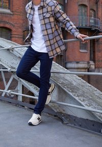 Camicia a scacchi beige e blu, t-shirt bianca, jeans scuri e sneakers beige con dettagli neri, in piedi su un parapetto di metallo.