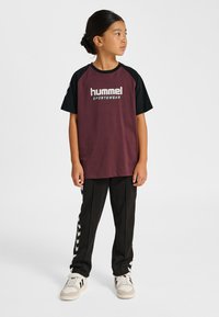 Vinröd och svart T-shirt med korta ärmar och "hummel SPORTSWEAR"-logotyp, kombinerad med svarta byxor med vita chevronränder och sneakers.