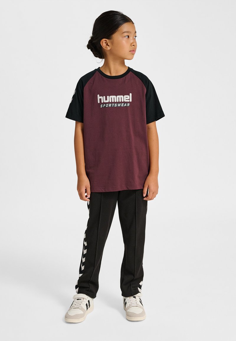 Vinröd och svart T-shirt med korta ärmar och "hummel SPORTSWEAR"-logotyp, kombinerad med svarta byxor med vita chevronränder och sneakers.