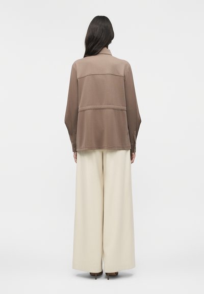 Γυναίκα με μακριά σκούρα μαλλιά, ντυμένη με φαρδιά καφέ ζακέτα και εκρού παντελόνι φαρδιάς γραμμής, στέκεται με την πλάτη γυρισμένη σε λευκό φόντο.