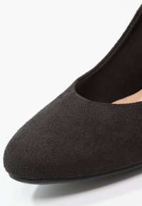 Gros plan sur une chaussure plate pour femme en daim noir avec un bout arrondi et un petit talon sur fond blanc.