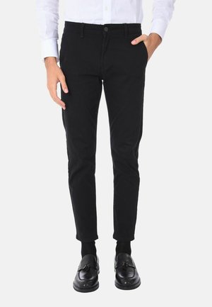 Pantaloni in cotone neri con vestibilità slim, dotati di chiusura a bottoni e tasche laterali, abbinati a mocassini neri e calze.