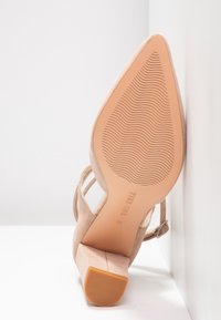 Chaussure à talons hauts pour femmes en daim beige, avec un motif ondulé sur la semelle et une bride à boucle, positionnée en angle sur une surface blanche devant un mur blanc.