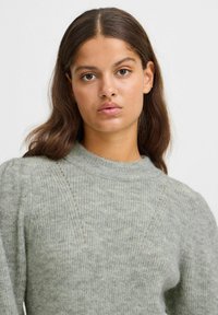 Pull en tricot vert clair avec un col haut, un jeu de textures en côtes et de subtils détails de coutures diagonales, offrant une apparence douce et confortable.