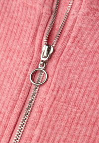 Vaaleanpunainen ribbineuloksinen kangas, jossa hopeanvärinen metallivetoketju. Vetoketjussa on pyöreä vetonuppi ja yksityiskohtaiset hammasosat.