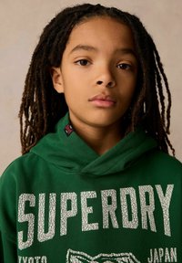 Grön hoodie med sprucken vit text "SUPERDRY" och logotyp. Har en huva med dragsko och svart etikett på kragen. Neutral bakgrund.