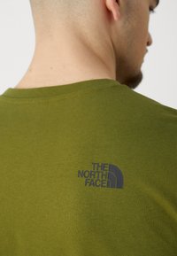 Olívazöld pamut póló kerek nyakkivágással, a hát felső részén a „THE NORTH FACE” fekete logójának nyomtatott változatával.