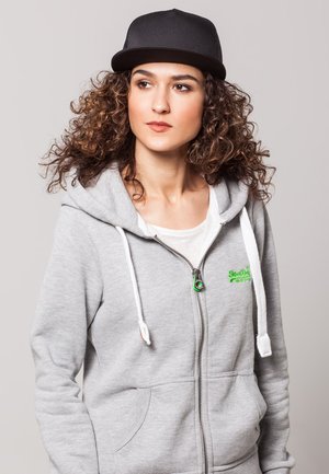 Grauer Zip-Hoodie mit weißen Kordelzügen und Fronttaschen, mit einem grünen Logo. Schwarze Flachschirmmütze. Lockiges Haar. Neutraler Hintergrund.