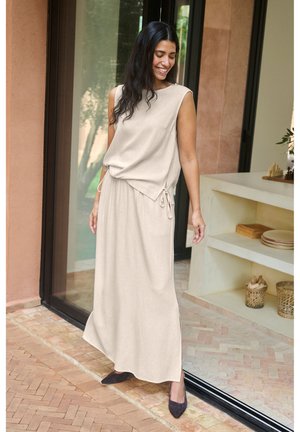 Donna sorridente in piedi vicino a una porta di vetro, che indossa un vestito beige senza maniche con legacci laterali e scarpe scure appuntite su pavimento piastrellato.