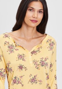 Langärmliges gelbes Oberteil mit floralen Druckmotiven in Pink und Grün. Verfügt über einen V-Ausschnitt mit Rüschensaum und einer fünf Knopfverschluss am Ausschnitt.