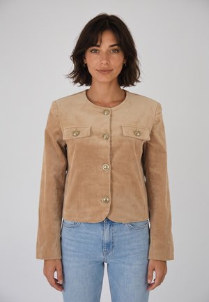 Mujer con cabello corto y oscuro que lleva una chaqueta de pana beige con botones y vaqueros azul claro contra un fondo claro y liso.