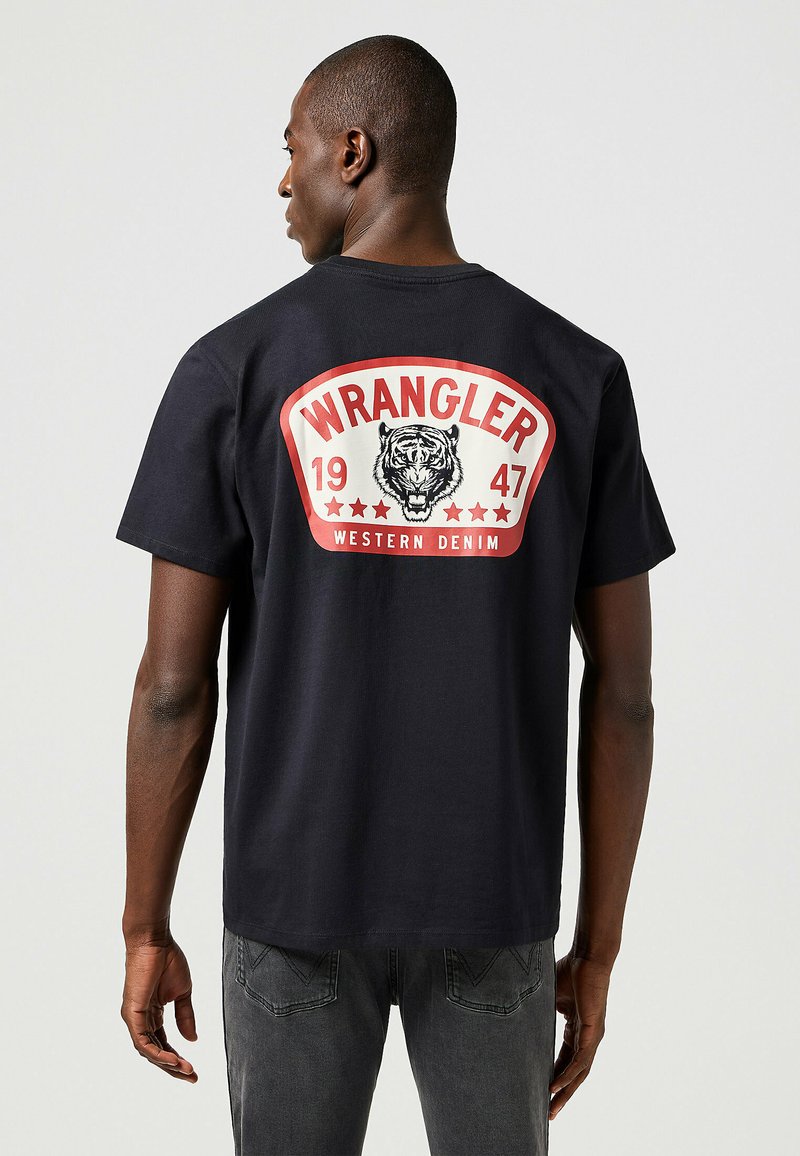 Camiseta de algodón negra con un gran gráfico rojo y blanco en la parte posterior que incluye el texto "WRANGLER", una ilustración de un tigre y estrellas.