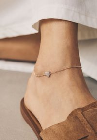 Goldene Fußkette mit einem herzförmigen Anhänger, der mit kleinen klaren Steinen verziert ist, getragen an einem Fuß mit einem hellbraunen Wildleder-Slipper.