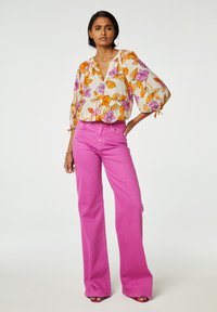 Bloemige blouse met witte achtergrond, oranje en paarse bloemen, en een V-hals. Roze wijde broek. Glad materiaal met een ontspannen pasvorm.