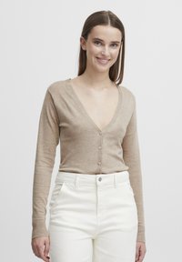 Cardigan beige en tricot léger et doux avec un décolleté en V profond, des boutons à l'avant et des manches longues, associé à un pantalon blanc taille haute.
