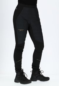 Schwarze isolierte Performance-Hosen mit einer schlanken Passform, die gepolsterte Abschnitte, goldene Akzente und mit Reißverschlüssen versehene Taschen haben. Kombiniert mit schwarzen Wanderschuhen.