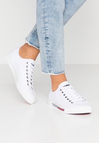 Person trägt hellblaue Jeans mit ausgefranstem Saum und weiße Canvas-Sneaker mit niedrigem Schaft und rot-weiß-blauer Streifenverzierung an der Sohle.