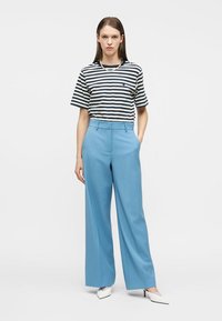 Γυναίκα που φοράει μπλούζα με κοντά μανίκια σε μαύρο και άσπρο ριγέ σχέδιο, ανοιχτό μπλε παντελόνι φαρδύ, λευκά μυτερά τακούνια, και στέκεται με το ένα χέρι στην τσέπη.