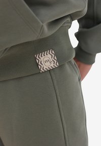 Oliwkowe dresy mają prążkowany pas z kwadratową metką, na której znajduje się falisty czarno-biały wzór oraz tekst "ja."