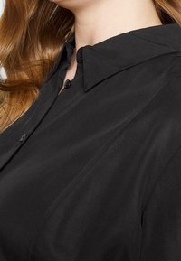 Schwarze Bluse mit klassischem Kragen, Knopfleiste und glattem Stoff mit subtilen Steppdetails.