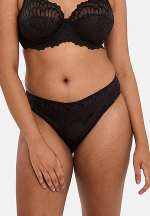 Soutien-gorge en dentelle noire transparente et culotte taille haute assortie portés sur la taille d'une personne au teint moyen.