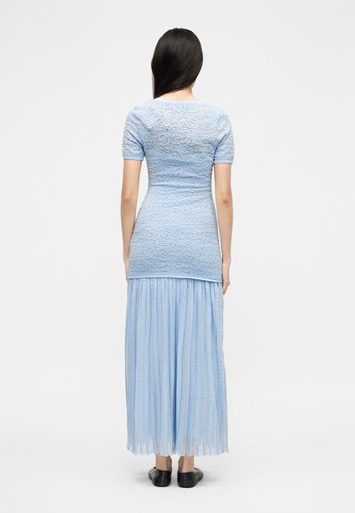 Γυναίκα με μακριά σκούρα μαλλιά, φορώντας ανοιχτό μπλε δαντελωτό κοντομάνικο τοπ και πλισέ μακριά φούστα, στέκεται ξυπόλητη μπροστά σε λευκό φόντο.