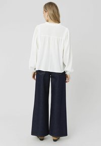 Blouse blanche texturée avec des manches bouffantes et un col à volants, associée à un jean large bleu foncé. La vue de dos montre un denim évasé et un minimum de détails.