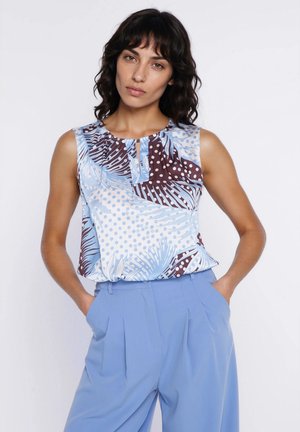 Kobieta w bezrękawniku w wzory w kolorach niebieskim, białym i brązowym, w wysokich jasnoniebieskich spodniach z rękami w kieszeniach.