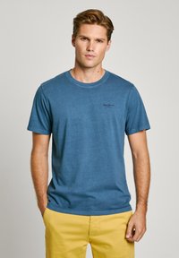 Μπλε βαμβακερό t-shirt με στρογγυλή λαιμόκοψη και κοντά μανίκια. Διαθέτει μικρό μαύρο λογότυπο στο στήθος. Συνδυάζεται με κίτρινες βερμούδες.