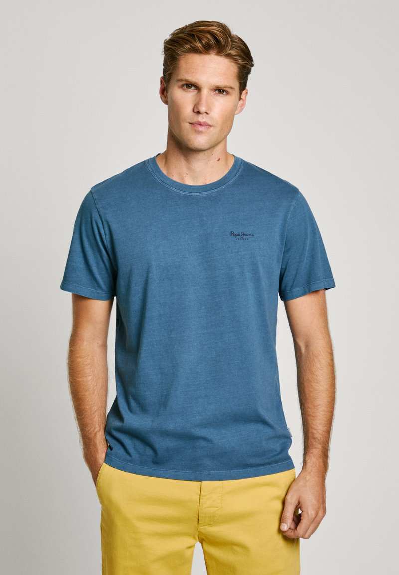 Μπλε βαμβακερό t-shirt με στρογγυλή λαιμόκοψη και κοντά μανίκια. Διαθέτει μικρό μαύρο λογότυπο στο στήθος. Συνδυάζεται με κίτρινες βερμούδες.