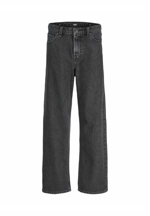Personne portant une veste beige, une chemise à carreaux bleue en dessous, un jean gris foncé ample et des chaussures beiges à lacets, debout devant un fond uni.