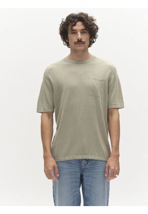 Mężczyzna o kręconych włosach i wąsach, ubrany w beżową koszulę z krótkim rękawem z przednią kieszenią oraz jasnoniebieskie dżinsy, stojący na jednolitym białym tle.