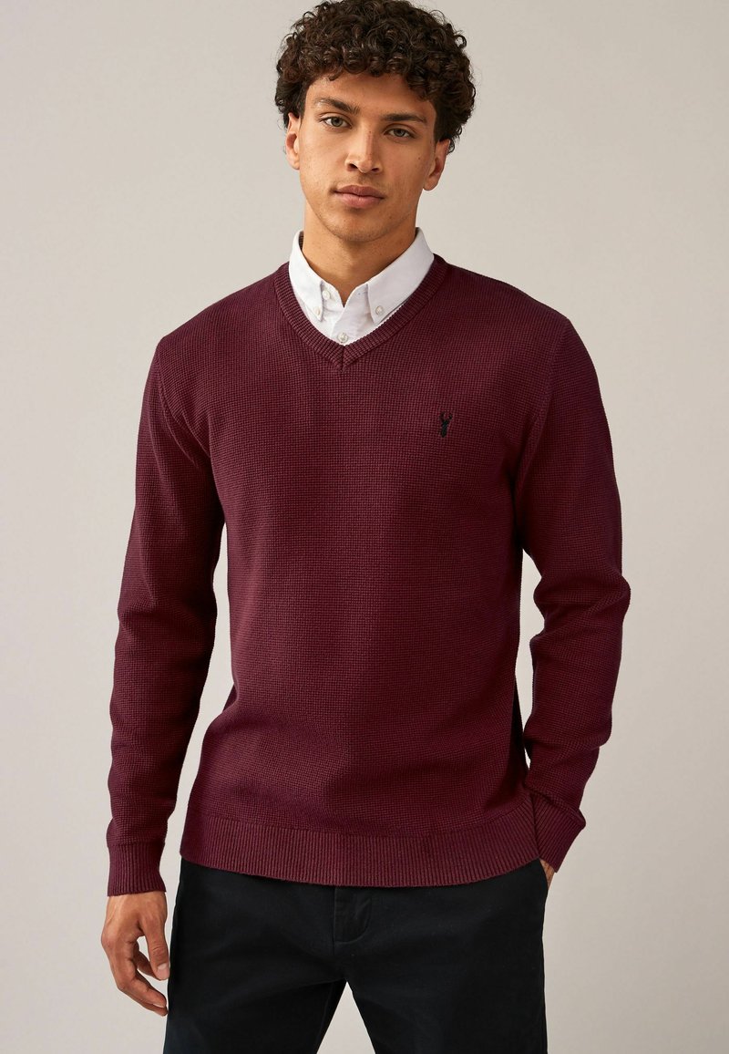 Pull en v burgundy avec un motif tricoté texturé, des poignets et un ourlet côtelés. Présente un petit logo brodé sur la poitrine.