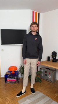 Felpa nera con logo rosso, pantaloni beige, scarpe slip-on nere e calzini bianchi. In piedi in una stanza moderna con una TV, piante e una lampada colorata.