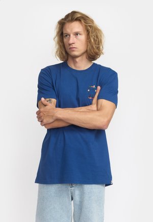 Blå bomullströja med kort ärm, med en liten flerfärgad grafisk fickdesign. Bärs med ljusa denimjeans.
