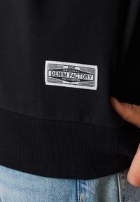 Felpa nera in tessuto con polsini e fondo a costine, dotata di un'etichetta bianca che recita "Original Denim Factory" sul lato sinistro.