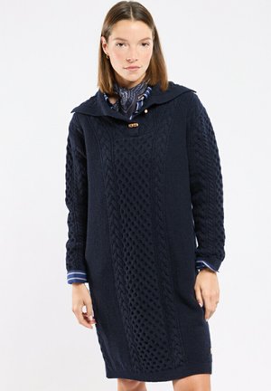 Femme portant une robe-pull en maille torsadée bleu marine avec capuche et bouton à bascule sur une chemise rayée et un foulard à motifs autour du cou.
