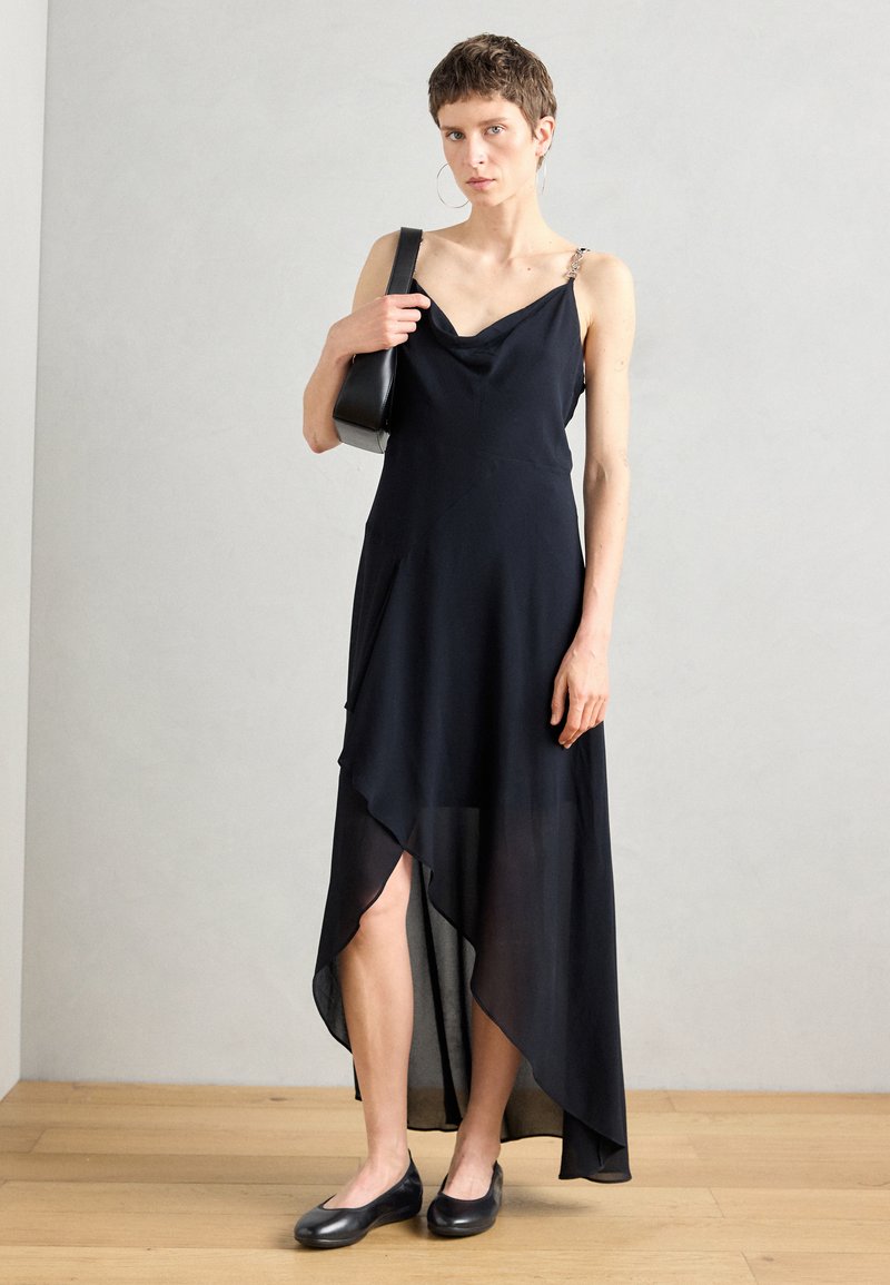 Schwarzes asymmetrisches Kleid mit einem schalenförmigen Ausschnitt, durchsichtigem Stoff und mit Kettenakzenten versehenen Trägern. Kombiniert mit schwarzen Ballerinas und einer kleinen schwarzen Tasche.