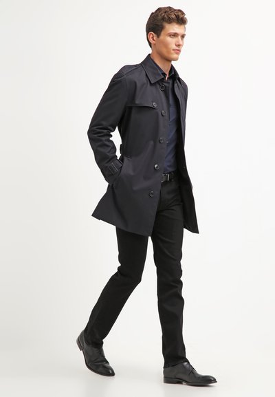 Manteau trench en marine avec un col structuré, boutons double boutonnage, poches latérales, porté sur une chemise sombre et un pantalon slim, associé à des chaussures noires.