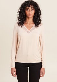 Beige Langarmoberteil mit einem Spitzen-V-Ausschnitt, aus weichem Stoff und mit lockerer Passform, kombiniert mit schwarzen Hosen.