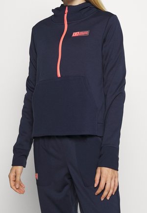 Kvinde iført navyblåt hættetrøje- og bukse sæt med lys koral lynlås og mærkede patches, stående mod en ensfarvet lys baggrund.