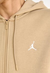 Beżowa bluza na zamek z miękkiego materiału z kieszenią kangurka, z białym logo Jumpman na lewej piersi i kapturem ze sznurkiem.