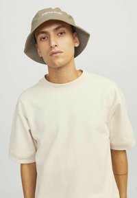 Beige bucket hat with a wide brim and embroidered “Original Studio” logo. Worn with a light-colored, oversized T-shirt.