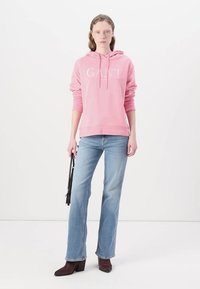 Rosa bomullshuvtröja med ordet "GANT" i vit text, ihop med ljusblå utsvängda jeans och bruna klackstövlar, hållandes med en svart väska.